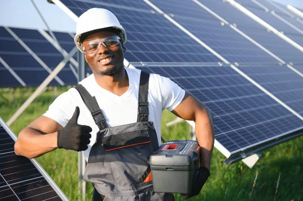 Koruyucu miğfer takmış yakışıklı Afrikalı Amerikalı zanaatkârın portresi. Üniformalı ve aletleri güneş panellerinin arasında duran bir adam..