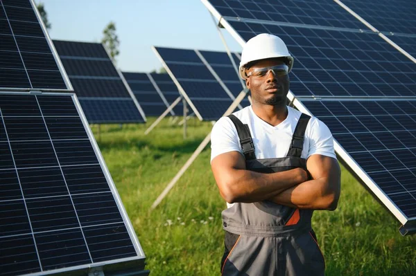 Beyaz kasklı ve gri tulumlu Afro-Amerikan adam güneş panellerinin arasında duruyor..