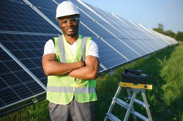Koruyucu miğfer takmış yakışıklı Afrikalı Amerikalı zanaatkârın portresi. Üniformalı ve aletleri güneş panellerinin arasında duran bir adam..