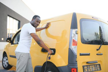 Afrikalı bir adam elektrik kablosunu elinde tutuyor. Elektrikli arabanın yanında duruyor..