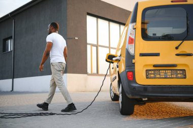 Elektrikli taşıt şarj istasyonlarında Afro-Amerikalı bir adam sarı elektrik dağıtım aracının yanında duruyor..