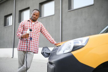 Afrikalı bir adam elektrik kablosunu elinde tutuyor. Elektrikli arabanın yanında duruyor..