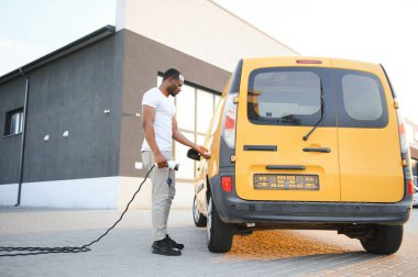 Elektrikli taşıt şarj istasyonunda Afrikalı Amerikalı adam elektrik dağıtım kamyonlarının yanında duruyor..