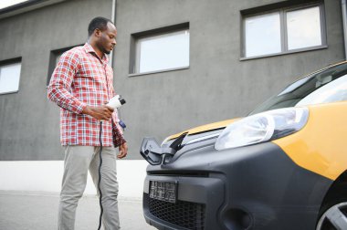 Afrikalı bir adam elektrik kablosunu elinde tutuyor. Elektrikli arabanın yanında duruyor.