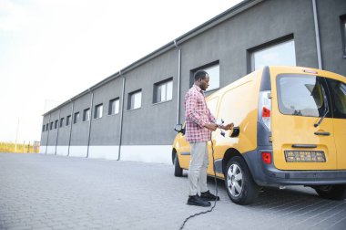 Elektrikli taşıt şarj istasyonunda Afrikalı Amerikalı adam elektrik dağıtım kamyonlarının yanında duruyor..