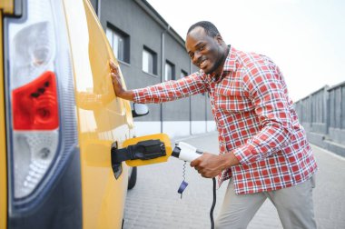 Afrikalı bir adam elektrik kablosunu elinde tutuyor. Elektrikli arabanın yanında duruyor.