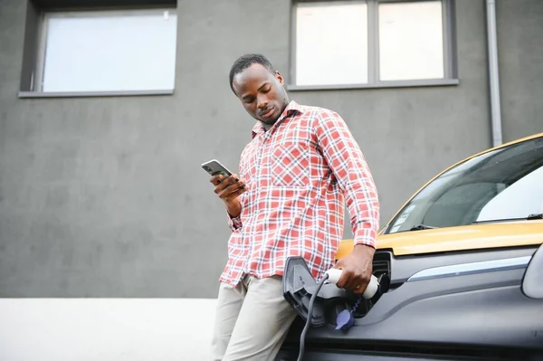 Homem Africano Casual Com Smartphone Perto Carro Elétrico Esperando Acabamento — Fotografia de Stock