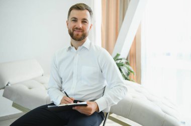 Terapi sırasında kameraya gülümseyen dost canlısı Orta Doğulu erkek psikolog klinikte çalışıyor, boş yer var. Ofiste poz veren mutlu psikoterapist