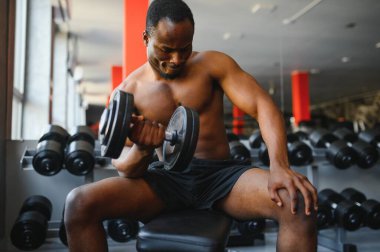 Genç Afrikalı Amerikalı bir adam spor salonundaki rafa yakın bir yerde oturuyor ve dambılı kaldırıyor. Vücut geliştirme merkezinde biceps kıvıran erkek ağırlık eğitmeni..