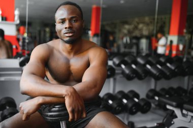 Seksi Afrikalı Amerikalı genç vücut geliştirici spor salonunda halter kaldırıyor, kolları üzerinde çalışıyor, fotokopi alanına bakıyor. Siyah, kaslı, üstsüz adam pazı egzersizi yapıyor. Sağlıklı yaşam tarzı kavramı