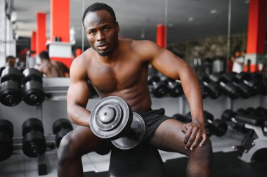 Genç Afrikalı Amerikalı bir adam spor salonundaki rafa yakın bir yerde oturuyor ve dambılı kaldırıyor. Vücut geliştirme merkezinde biceps kıvıran erkek ağırlık eğitmeni..