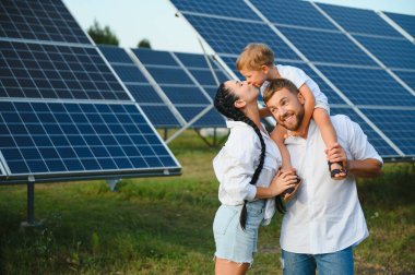 Üç kişilik genç aile fotovoltaik güneş panelinin yanında çömeliyor, küçük çocuk ve ebeveynler. Modern aile kavramı. Yeşil enerji kavramı.