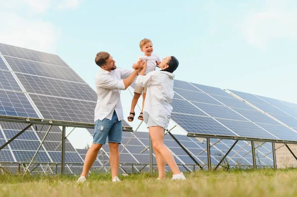 A wide shot of a happy family standing together and smiling at camera with a large solar panel in background.