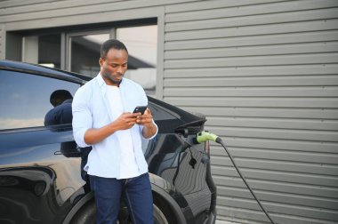 Afrikalı adam elektrik kablosuyla elektrikli arabanın yanında duruyor. Çevre dostu araç istasyonda şarj oluyor. Yaşam Tarzı konsepti.