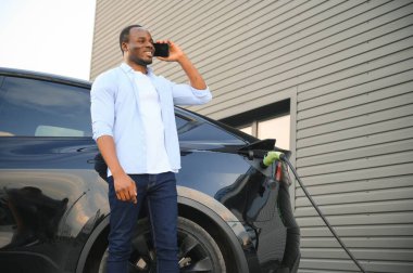 Elektrikli araç şarj istasyonunda cep telefonu olan mutlu genç adam..