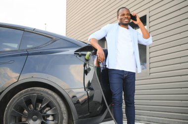 Afrikalı bir adam elektrikli bir arabanın yanında duruyor şarj istasyonunda şarj olmasını bekliyor ve telefonu kullanıyor.