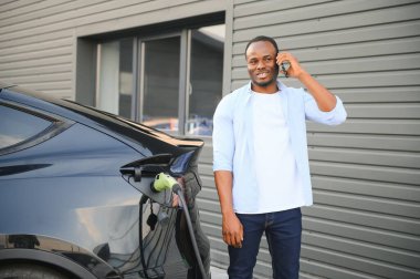 Elektrikli araç şarj istasyonunda cep telefonu olan mutlu genç adam..