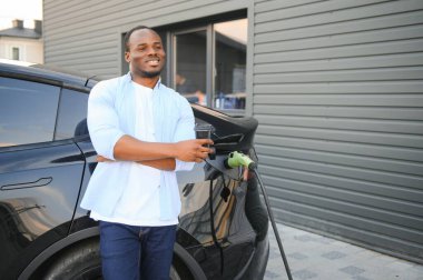 Afrikalı adam elektrik kablosuyla elektrikli arabanın yanında duruyor. Çevre dostu araç istasyonda şarj oluyor. Yaşam Tarzı konsepti.