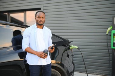 Afrikalı bir adam elektrikli bir arabanın yanında duruyor şarj istasyonunda şarj olmasını bekliyor ve telefonu kullanıyor.