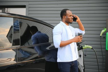 Afro-Amerikalı bir adam elektrikli bir arabanın yanında duruyor şarj istasyonunda şarj olmasını bekliyor ve kahve içiyor.