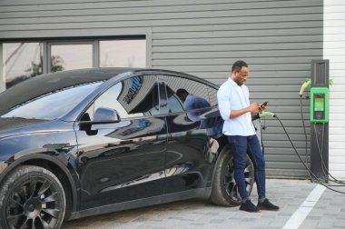 Afrikalı adam elektrik kablosuyla elektrikli arabanın yanında duruyor. Çevre dostu araç istasyonda şarj oluyor. Yaşam Tarzı konsepti.