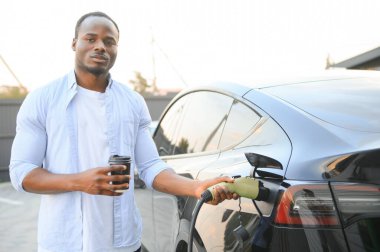 Afro-Amerikalı bir adam elektrikli bir arabanın yanında duruyor şarj istasyonunda şarj olmasını bekliyor ve kahve içiyor.