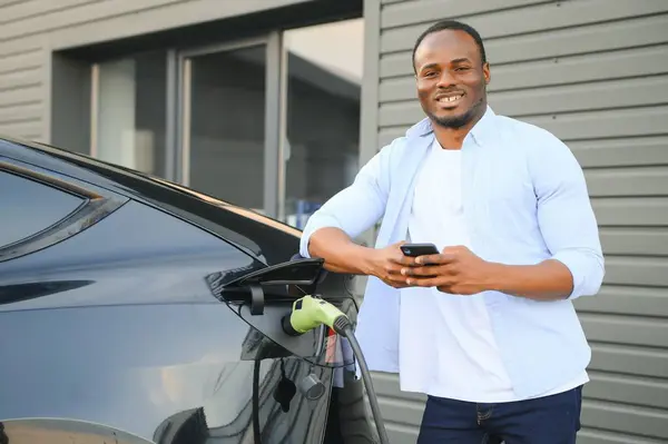 Happy young man with mobile phone charging car at electric vehicle charging station.