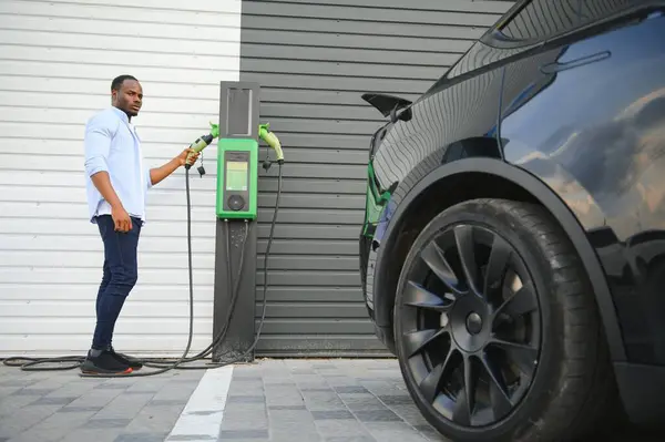高級電気自動車の近くに立って手元に充電ケーブルを持っている深刻なアフリカ人男性 — ストック写真