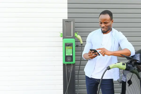 Happy young man with mobile phone charging car at electric vehicle charging station.