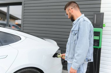 Güzel, genç, şık bir adam, gündüz vakti elektrikli arabasıyla aracı şarj ediyor..