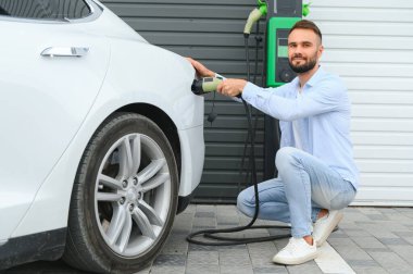 Genç yakışıklı adam elektrik şarj istasyonunda kablo tutuyor. Yeni arabasının yanında duruyor..