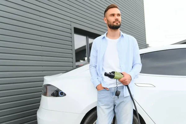 Man Holding Power Charging Cable For Electric Car In Outdoor Car Park. And he s going to connect the car to the charging station in the parking lot near the shopping center