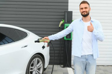 Güzel, genç, şık bir adam, gündüz vakti elektrikli arabasıyla aracı şarj ediyor..
