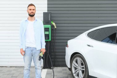 Güzel, genç, şık bir adam, gündüz vakti elektrikli arabasıyla aracı şarj ediyor..