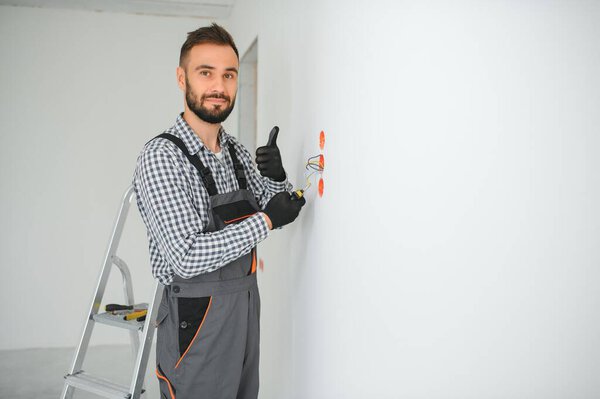 Electrician in uniform mounting electric sockets on the white wall indoors.