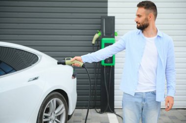 Genç yakışıklı adam elektrik şarj istasyonunda kablo tutuyor. Yeni arabasının yanında duruyor..