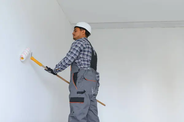 An Indian apartment repair worker paints a white wall with a roller