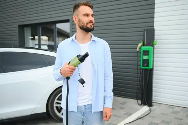 Man Holding Power Charging Cable For Electric Car In Outdoor Car Park. And he s going to connect the car to the charging station in the parking lot near the shopping center