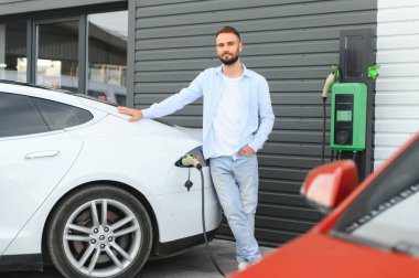 Güzel, genç, şık bir adam, gündüz vakti elektrikli arabasıyla aracı şarj ediyor..