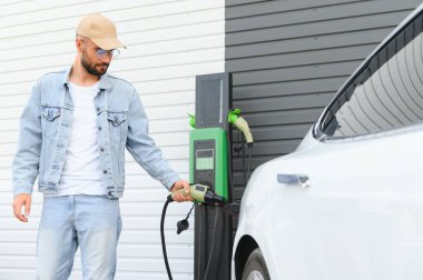 Genç yakışıklı adam elektrik şarj istasyonunda kablo tutuyor. Yeni arabasının yanında duruyor..