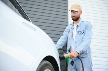 Güzel, genç, şık bir adam, gündüz vakti elektrikli arabasıyla aracı şarj ediyor..