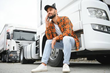 Aracın yanına yaslanmış ve telefon kullanıyor. Genç kamyon şoförü gündüz vakti..