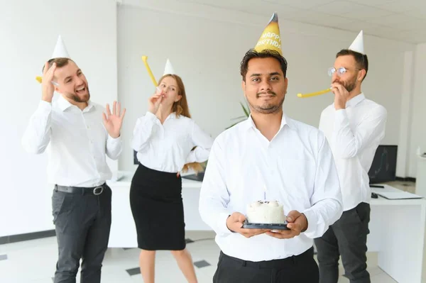 happy business team with birthday cake and gifts greeting indian male colleague at office party.