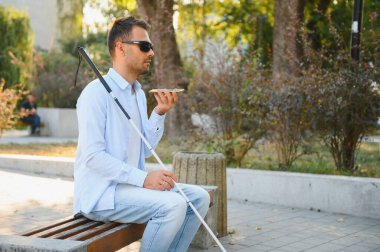 Elinde baston olan kör bir adam bankta oturur ve arka planda akıllı telefon kullanır.