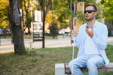 Elinde baston olan kör bir adam bankta oturur ve arka planda akıllı telefon kullanır.