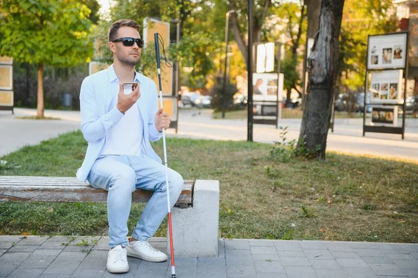 ベンチに座っている歩く棒を持つ盲目の人は 都市の背景にスマートフォンを使用します ストック写真