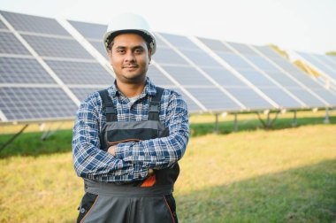 Mavi gökyüzüne karşı güneş panellerinin yanında duran beyaz şapkalı genç Hintli teknisyen portresi. Endüstriyel işçi güneş sistemi kurulumu, yenilenebilir yeşil enerji üretimi kavramı