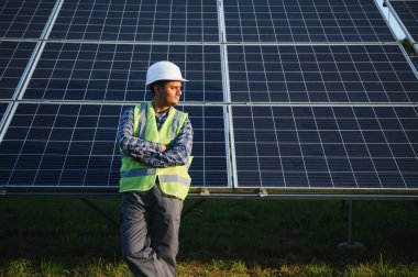 Mavi gökyüzüne karşı güneş panellerinin yanında duran beyaz şapkalı genç Hintli teknisyen portresi. Endüstriyel işçi güneş sistemi kurulumu, yenilenebilir yeşil enerji üretimi kavramı