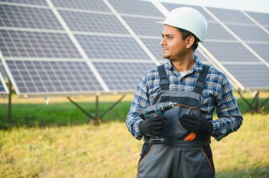 Mavi gökyüzüne karşı güneş panellerinin yanında duran beyaz şapkalı genç Hintli teknisyen portresi. Endüstriyel işçi güneş sistemi kurulumu, yenilenebilir yeşil enerji üretimi kavramı
