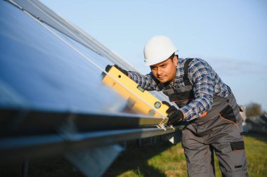 Mavi gökyüzüne karşı güneş panellerinin yanında duran beyaz şapkalı genç Hintli teknisyen portresi. Endüstriyel işçi güneş sistemi kurulumu, yenilenebilir yeşil enerji üretimi kavramı
